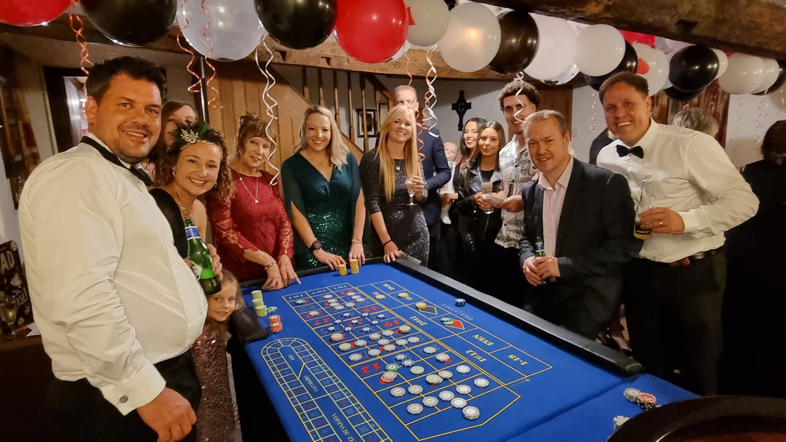 Guests in black tie around a fun casino roulette table at a party decorated with red, black and white balloons